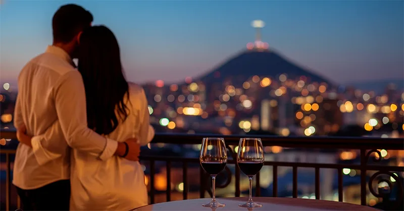 Pareja en una noche de pasión en uno de los mejores moteles de Cali, disfrutando la vista de la ciudad y Cristo Rey con copas de vino.