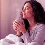 Mujer joven relajada y sonriendo plácidamente en la cama con una taza de café, representando la conexión entre el bienestar sexual, la reducción del estrés y la salud mental.