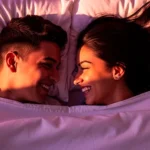 Pareja joven colombiana en la cama, sonriendo y mirándose a los ojos con complicidad bajo un edredón blanco. Representa la importancia de la conexión emocional y la calidad en la vida sexual en Colombia, según la encuesta de Forbes.