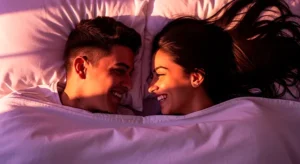 Pareja joven colombiana en la cama, sonriendo y mirándose a los ojos con complicidad bajo un edredón blanco. Representa la importancia de la conexión emocional y la calidad en la vida sexual en Colombia, según la encuesta de Forbes.
